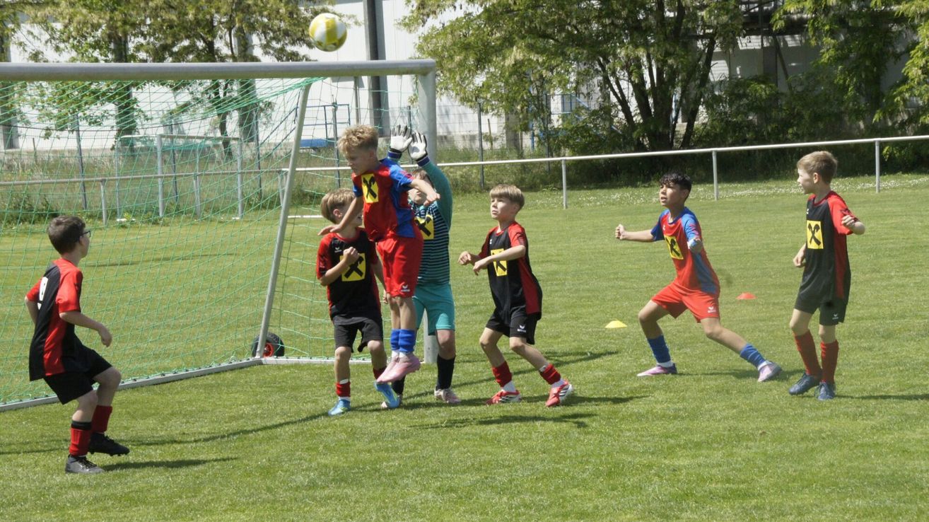 Fünf Kinder spielen Fußball auf einem Feld mit einem Tor. Ein Kind springt, um den Ball zu blockieren, während ein anderes auf den Ball zuläuft. Die anderen drei Kinder laufen auf den Ball zu.