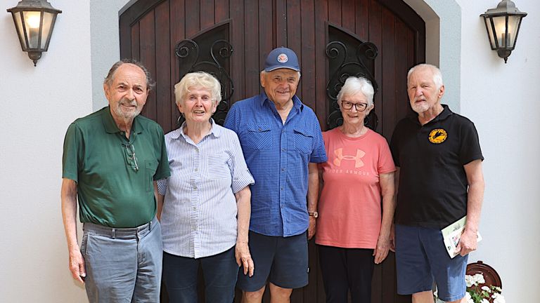 Fünf Menschen stehen lächelnd vor einer Holztür. Ein Mann trägt eine Kappe, ein anderer ein rosa T-Shirt und ein dritter ein schwarzes Shirt.