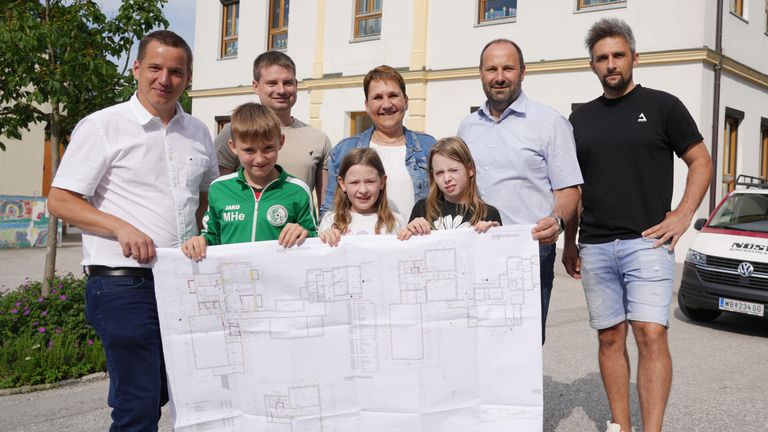 Eine Gruppe von Menschen steht vor einem Gebäude, lächelt und hält einen großen Bauplan. Dazu gehören zwei Frauen und vier Kinder, von denen eines ein grünes Jackett trägt.
