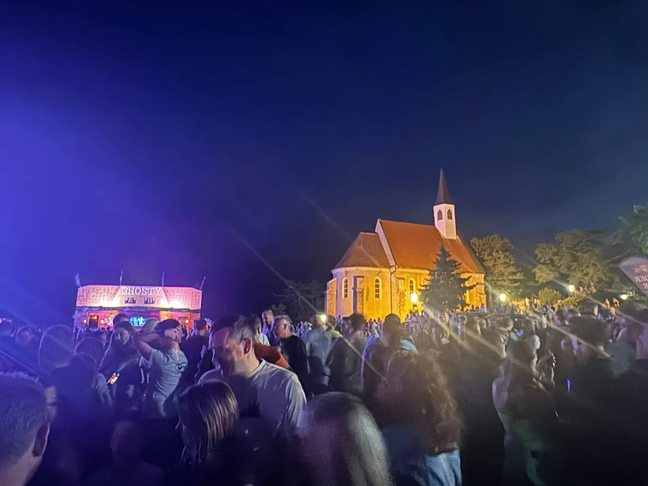 Eine Menschenmenge versammelt sich bei Nacht vor einer Kirche, die von lila Lichtern beleuchtet wird, mit Bäumen und einem Gebäude mit einem Schild, das 'GHOSTY' im Hintergrund liest.
