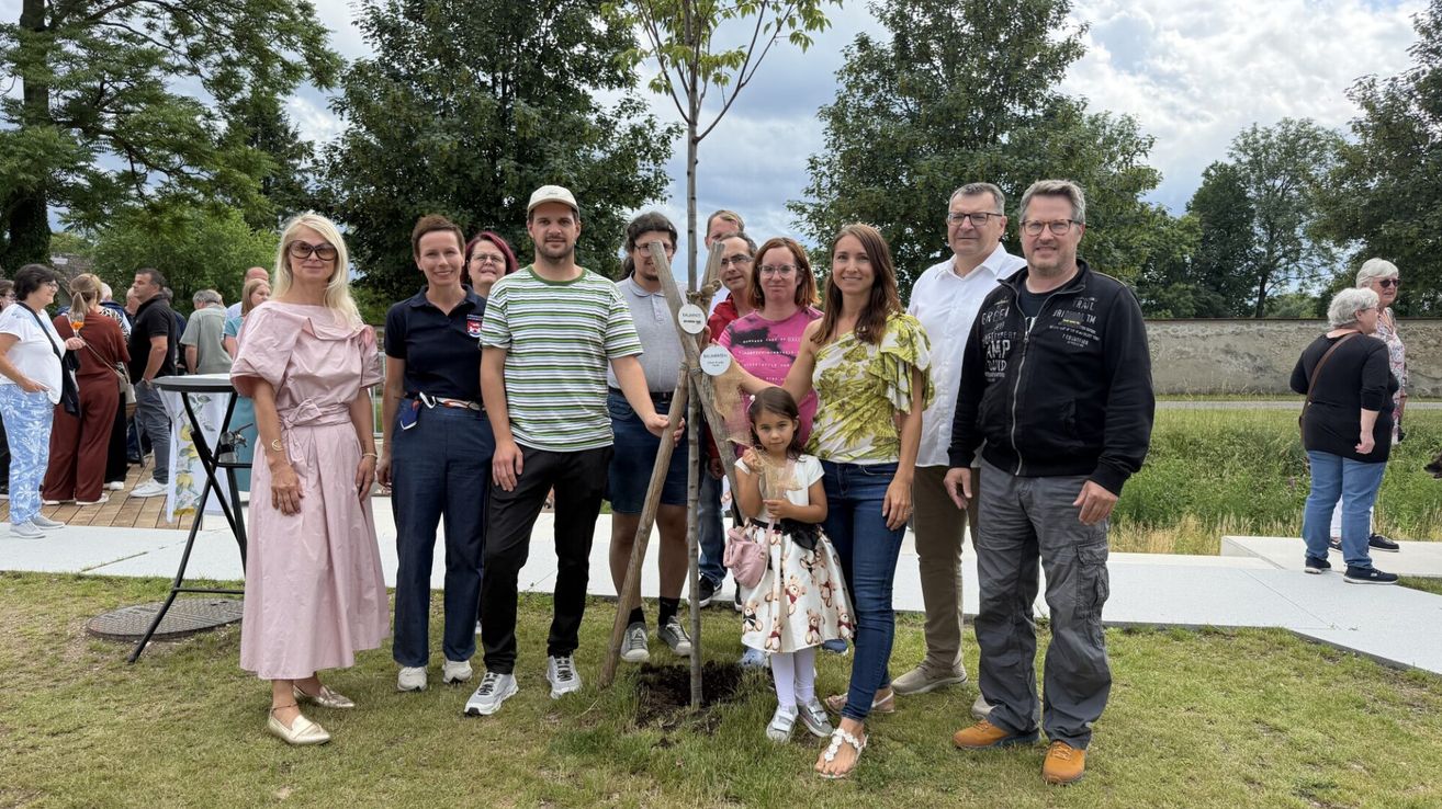 Eine Gruppe von Menschen steht um einen frisch gepflanzten Baum und lächelt für ein Foto. Einige tragen Brillen und Turnschuhe. Ein kleines Mädchen hält ein kleines Objekt in der Hand.