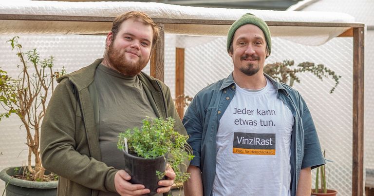 Zwei Männer lächeln in einem Gewächshaus. Einer hält eine Topfpflanze, der andere trägt eine grüne Mütze und ein weißes T-Shirt mit einer Botschaft.