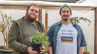 Zwei Männer lächeln in einem Gewächshaus. Einer hält eine Topfpflanze, der andere trägt eine grüne Mütze und ein weißes T-Shirt mit einer Botschaft.