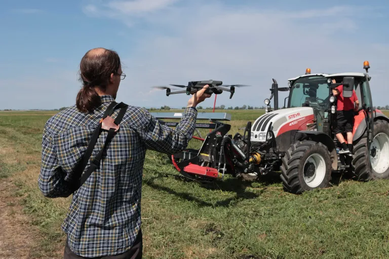 Eine Person mit Brille und kariertem Hemd steht in einem Feld, hält eine Drohne und schaut auf einen roten und weißen Traktor.