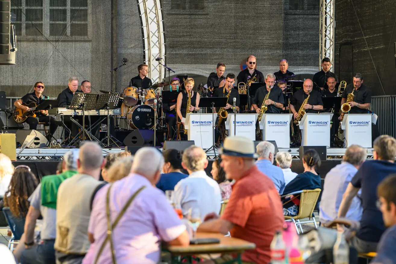 Eine Jazzband spielt auf einer Bühne mit einem Hintergrund, der mit Swingin' beschriftet ist. Die Musiker spielen Saxophone, Trompeten und andere Instrumente. Das Publikum sitzt davor und genießt die Aufführung.