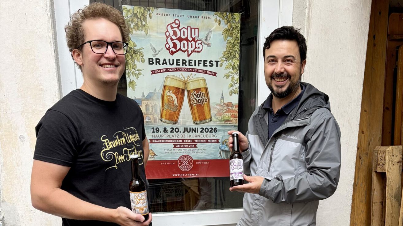 Zwei lächelnde Männer, einer in einem schwarzen T-Shirt und der andere in einer grauen Jacke, halten Bierflaschen vor einem Plakat, das für ein Bierfestival am 19. und 20. Juni wirbt.
