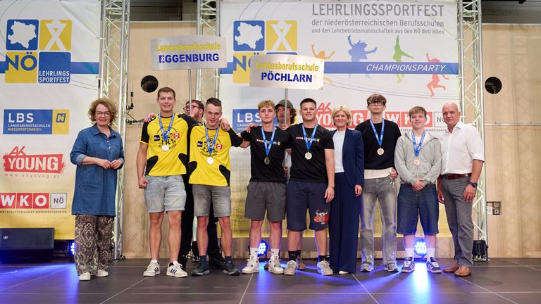 Fünf junge Männer mit Medaillen stehen auf einer Bühne. Dahinter stehen eine Frau und ein Mann. Ein Banner mit Text und Logos steht dahinter.