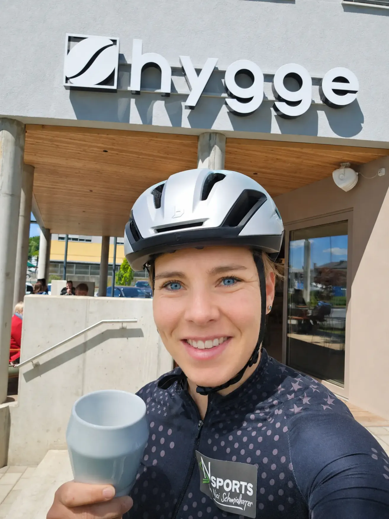 Eine lächelnde Frau in Fahrradausrüstung posiert vor einem Nyygge-Laden und hält eine Kaffeetasse. Der Hintergrund zeigt ein modernes Gebäude mit Glaswänden und einer Treppe.