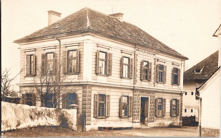 Ein altes Foto zeigt ein großes Gebäude mit vielen Fenstern und einem Schieferdach. Die Vorderseite trägt die Aufschrift Volkschule. Im Vordergrund links befindet sich eine Steinmauer und ein Baum.