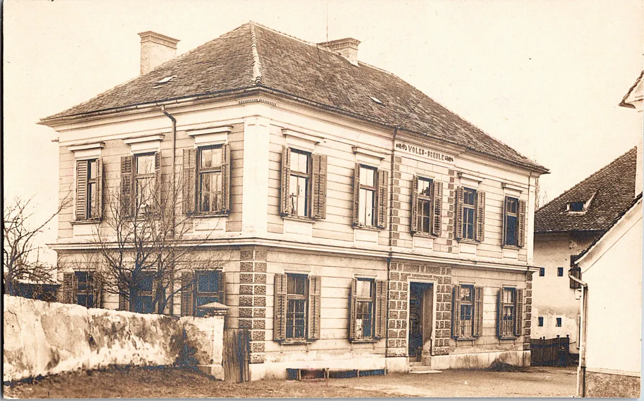 Ein altes Foto zeigt ein großes Gebäude mit vielen Fenstern und einem Schieferdach. Die Vorderseite trägt die Aufschrift Volkschule. Im Vordergrund links befindet sich eine Steinmauer und ein Baum.
