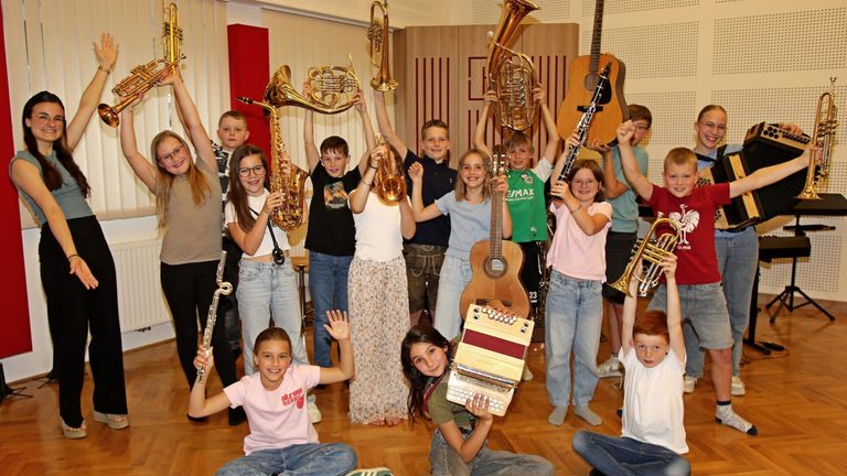 Eine Gruppe von Kindern in einem Musikraum, jeder hält verschiedene Musikinstrumente. Sie lächeln und posieren für ein Foto. Einige stehen, andere sitzen.