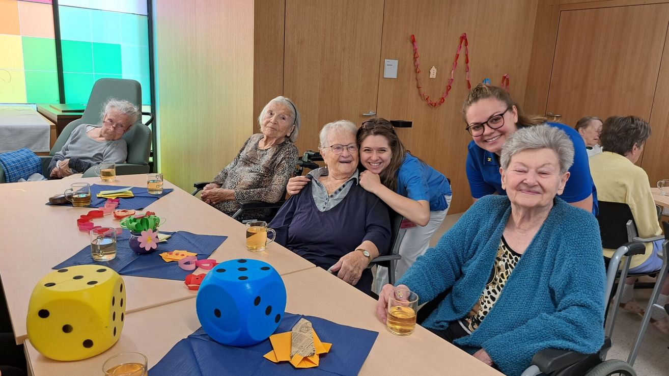 Eine Gruppe älterer Frauen sitzt an einem Tisch. Eine Frau ist im Rollstuhl, und eine andere umarmt sie. Auf dem Tisch liegt ein blauer Würfel, zusammen mit Getränkebechern und Servietten.