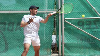 Ein Tennisspieler in einem weißen Shirt und Shorts schwingt einen Tennisschläger, möglicherweise während eines Matches. Er trägt eine schwarze Kappe und eine Armbanduhr. Hinter ihm steht ein grüner Zaun.