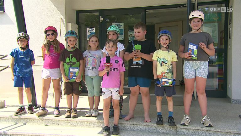 Eine Gruppe von Kindern mit Helmen steht vor einem Gebäude. Sie halten Mikrofone und Bücher. Die Kinder lächeln und scheinen für ein Foto zu posieren. Hinter ihnen ist eine Glastür mit einem Schild.