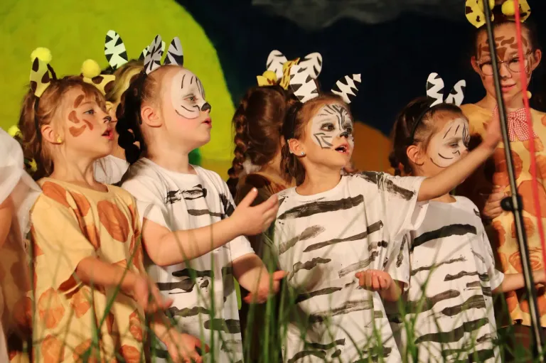 Eine Gruppe von Kindern in Tierkostümen und mit Gesichtsbemalung führt in einer Schulaufführung auf der Bühne auf.