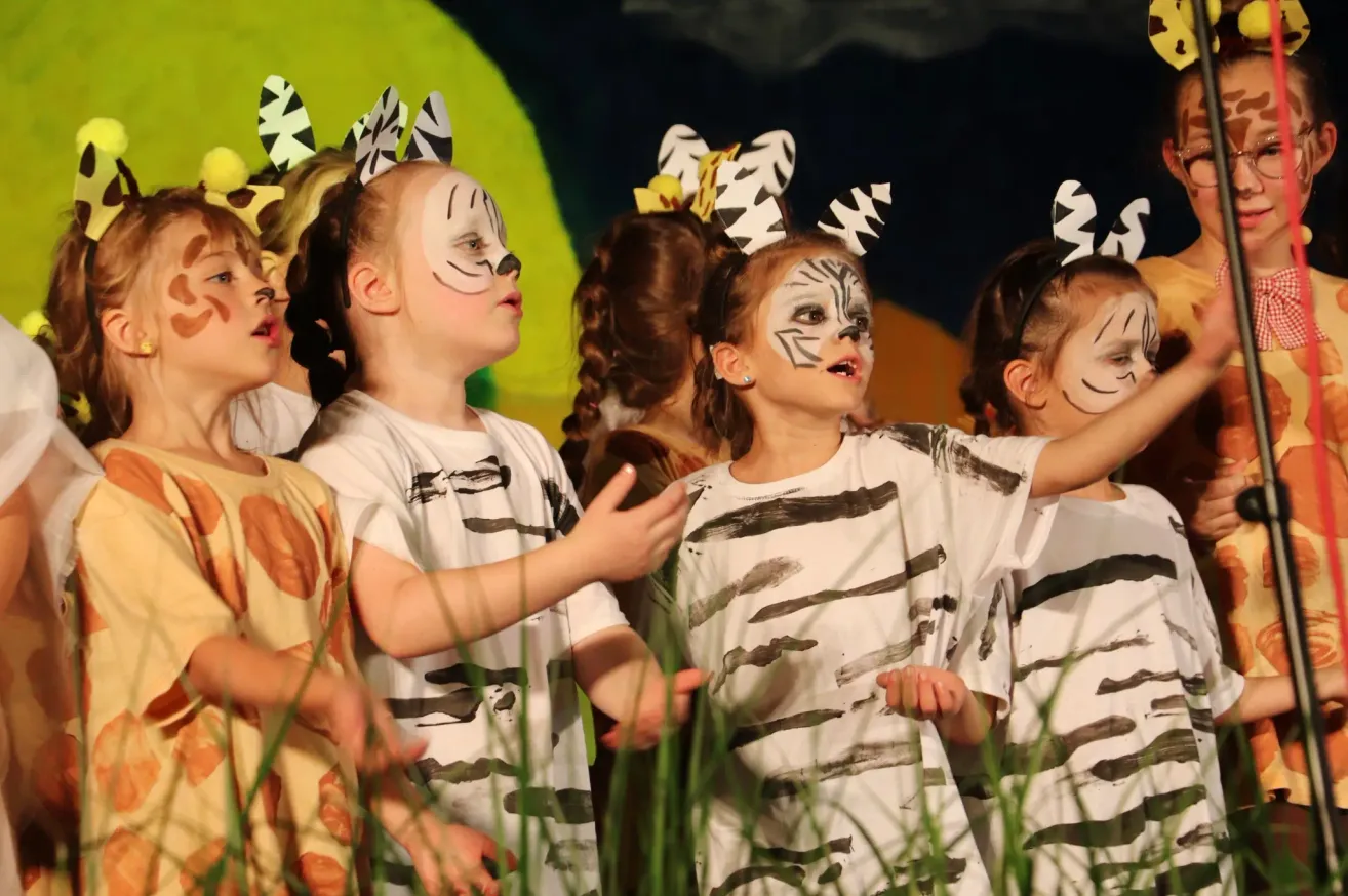 Eine Gruppe von Kindern in Tierkostümen und mit Gesichtsbemalung führt in einer Schulaufführung auf der Bühne auf.