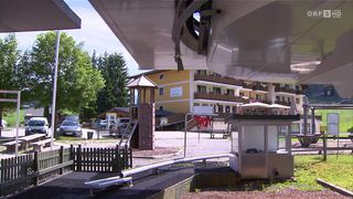 Ein Spielplatz mit Rutsche und Klettergerüst in der Nähe eines gelben Gebäudes mit mehreren Stockwerken und Balkonen. Im Hintergrund sind Bäume und geparkte Autos zu sehen.