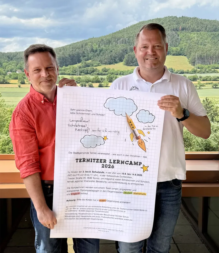 Zwei Männer halten ein Poster mit einer Rakete und Wolken. Einer trägt ein rotes Hemd, der andere ein weißes Poloshirt. Sie stehen vor einer malerischen Kulisse. Das Poster enthält eine Liste mit Details zu einem Lerncamp.