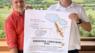 Zwei Männer halten ein Poster mit einer Rakete und Wolken. Einer trägt ein rotes Hemd, der andere ein weißes Poloshirt. Sie stehen vor einer malerischen Kulisse. Das Poster enthält eine Liste mit Details zu einem Lerncamp.