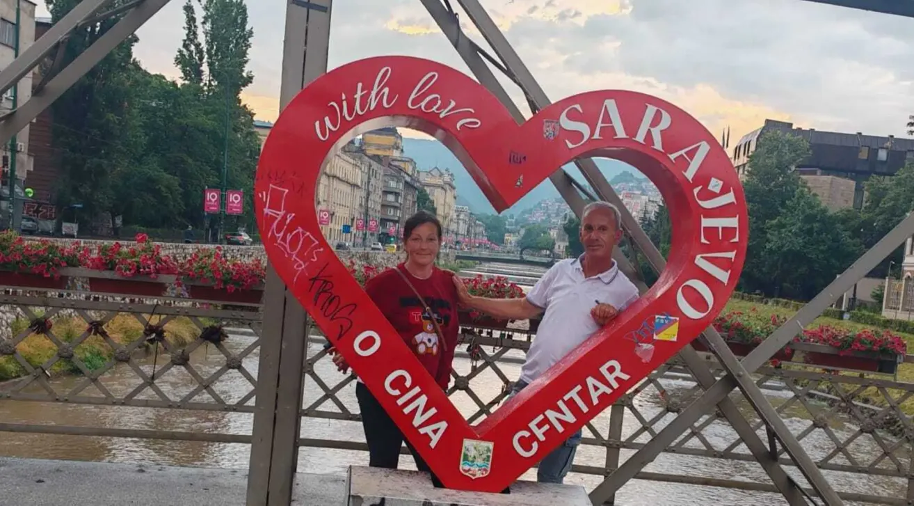 Ein Paar steht in einer roten herzförmigen Aussparung auf einer Brücke in Sarajevo und posiert für ein Foto. Die Frau trägt ein rotes Shirt, der Mann ein weißes.