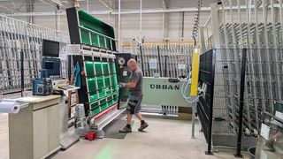 Ein Mann in einer Fabrik arbeitet an einer großen grünen Maschine. Die Maschine ist mit U-R-B-A-N beschriftet und hat einen großen grünen Bildschirm davor. Der Mann trägt Handschuhe und Turnschuhe.