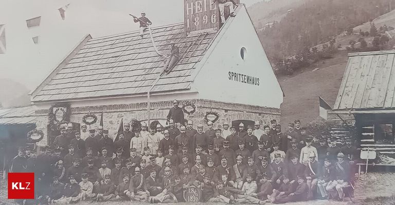 Ein historisches Foto von Feuerwehrleuten in Uniform vor einem Feuerwehrhaus mit dem Jahr 1896 auf dem Dach, zwei Männer auf dem Dach mit einem Seil und einer Leiter.
