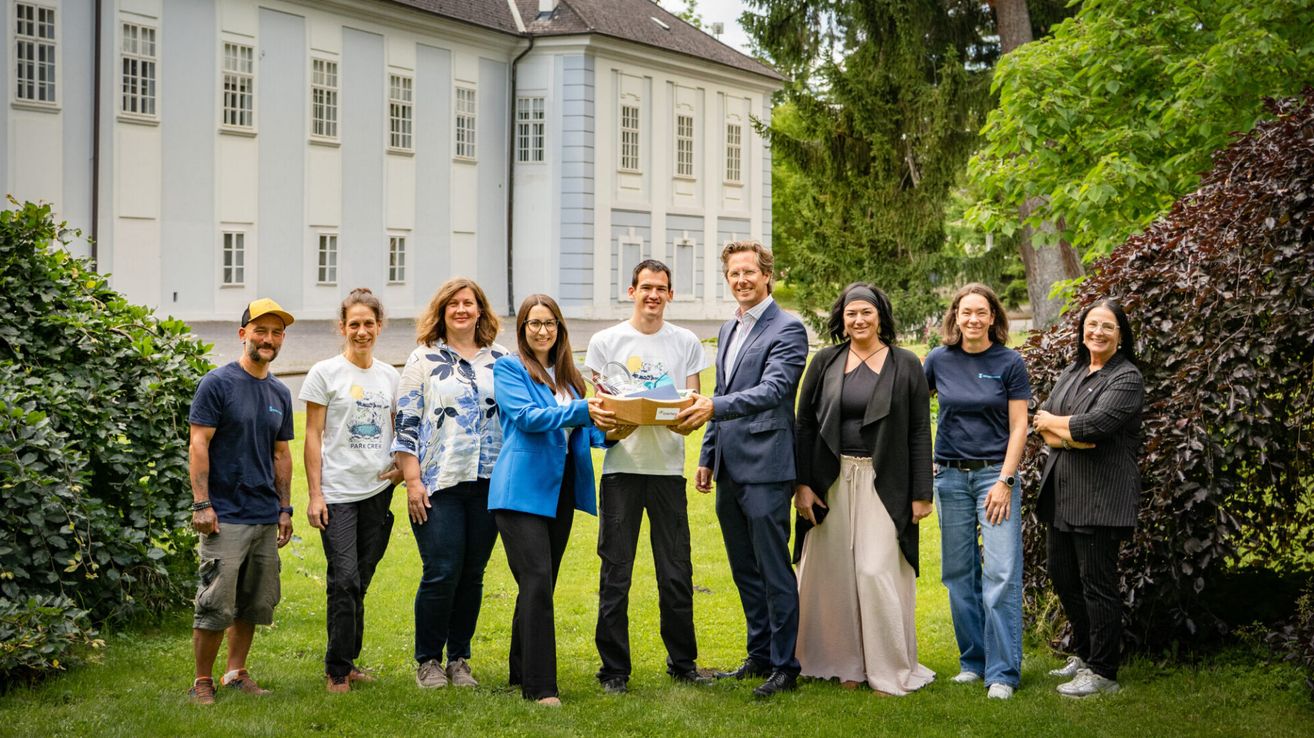 Eine Gruppe von Menschen steht vor einem Gebäude, lächelt und posiert für ein Foto. Ein Mann im Anzug überreicht einer Frau im blauen Mantel ein Geschenk.