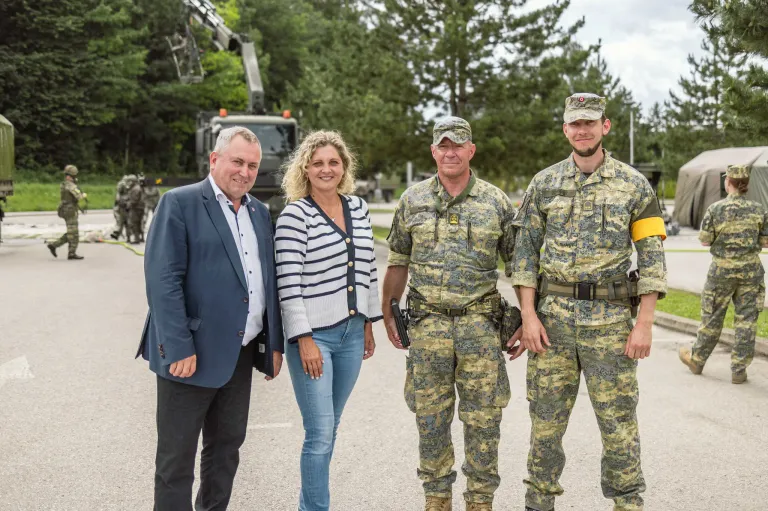 Drei Personen in Uniformen stehen vor einem Fahrzeug, eine Frau in einer Strickjacke und ein Mann in einem Anzug lächeln.