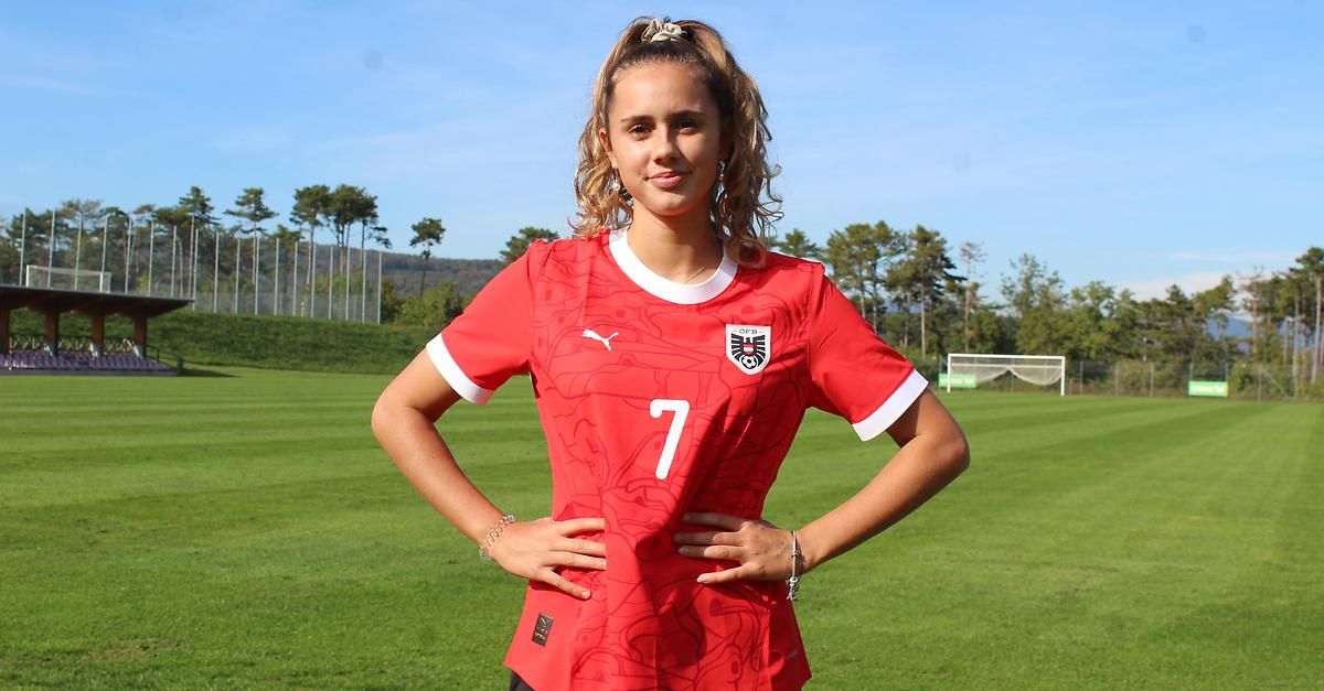 Eine Frau in einem roten Fußballtrikot mit der Nummer 7 posiert für ein Foto auf einem Feld. Im Hintergrund sind verschwommen Bäume und ein Fußballtor zu sehen.