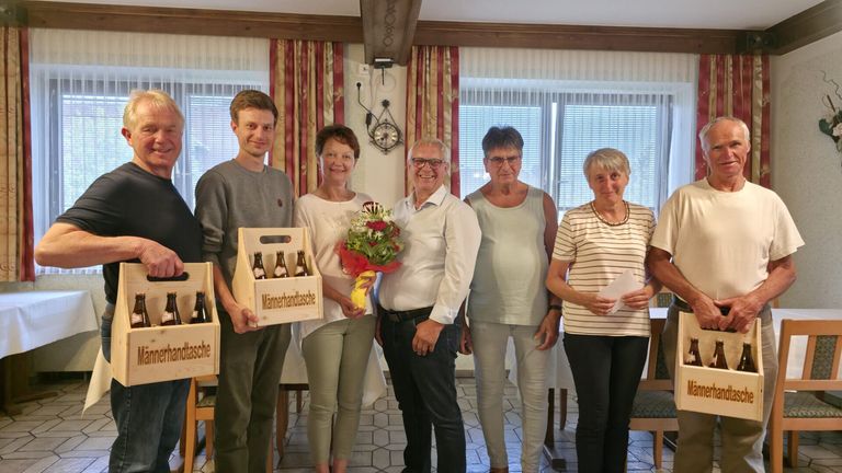 Sechs Menschen stehen in einem Raum, lächeln und posieren für ein Foto. Der Mann auf der linken Seite hält eine Holzkiste mit Flaschen. Der Mann in der Mitte hält einen Blumenstrauß. Die anderen halten Papiere. Hinter ihnen ist ein weißer Vorhang.