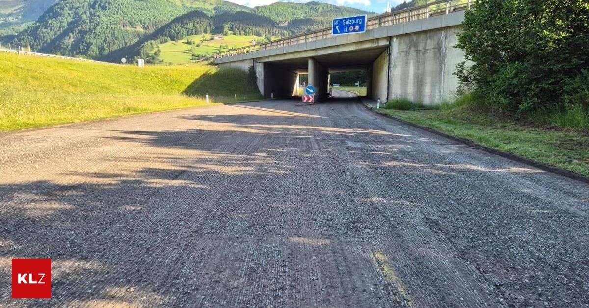 Eine leere Straße führt zu einem Tunnel unter einer Brücke. Im Hintergrund sind grünes Gras und Berge zu sehen. Ein Schild für Salzburg befindet sich über dem Tunnel.