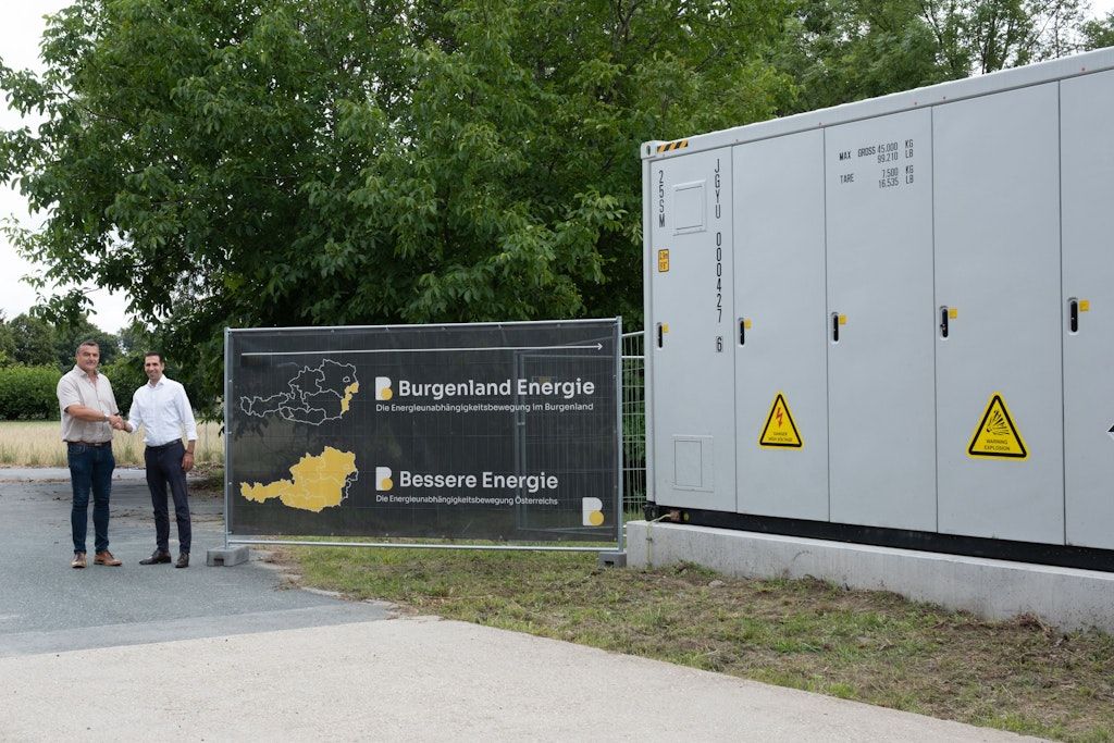 Ein Mann steht neben einem Schild für Burgenland Energie, ein gelbes und schwarzes Schild mit einem gelben Pfeil, der nach rechts zeigt. Der Hintergrund zeigt einen Baum und einen weißen Stromkasten.
