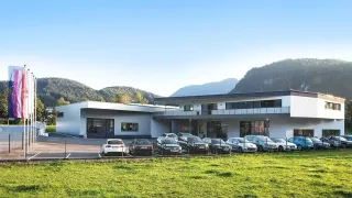 Ein großes Gebäude mit mehreren geparkten Autos davor, umgeben von einem grünen Rasen und einem Zaun. Im Hintergrund sind Berge sichtbar.