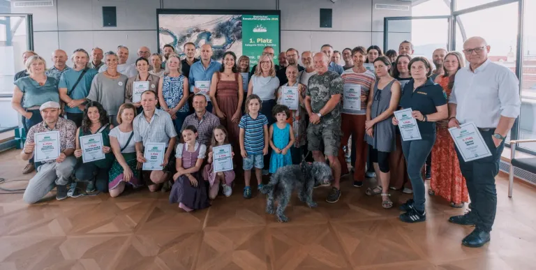 Eine Gruppe von Menschen, Kindern und einem Hund posiert mit Urkunden für ein Foto in einem Raum. Ein Banner hängt an der Wand.