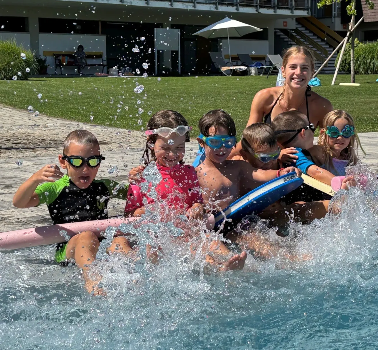 Eine Gruppe von Kindern und einer erwachsenen Frau spielen im Pool und tragen Schwimmbrillen. Sie spritzen Wasser und lächeln. Dahinter befindet sich eine Rasenfläche mit einem weißen Schirm und einigen Stühlen.