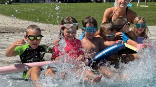Eine Gruppe von Kindern und einer erwachsenen Frau spielen im Pool und tragen Schwimmbrillen. Sie spritzen Wasser und lächeln. Dahinter befindet sich eine Rasenfläche mit einem weißen Schirm und einigen Stühlen.
