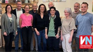 Eine Gruppe von Menschen, hauptsächlich Erwachsene, posiert für ein Foto in einem Raum. Sie lächeln alle und schauen in die Kamera. Sie tragen Freizeitkleidung. Hinter ihnen steht ein Holzregal mit einem weißen Objekt obenauf.