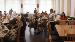 Eine Familie sitzt mit einem Baby auf dem Schoß an einem Tisch in einem Restaurant. Andere sind in der Nähe. Eine Frau steht vor ihnen und hält ein Glas.