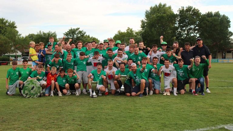 Eine Gruppe junger Männer in grünen Trikots und Mützen feiert einen Sieg auf einem Grasfeld, hält Trophäen und posiert für ein Foto.