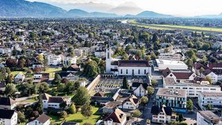 Luftaufnahme einer Stadt mit Gebäuden, einer Kirche und einem Fluss. In der Ferne sind Berge sichtbar.