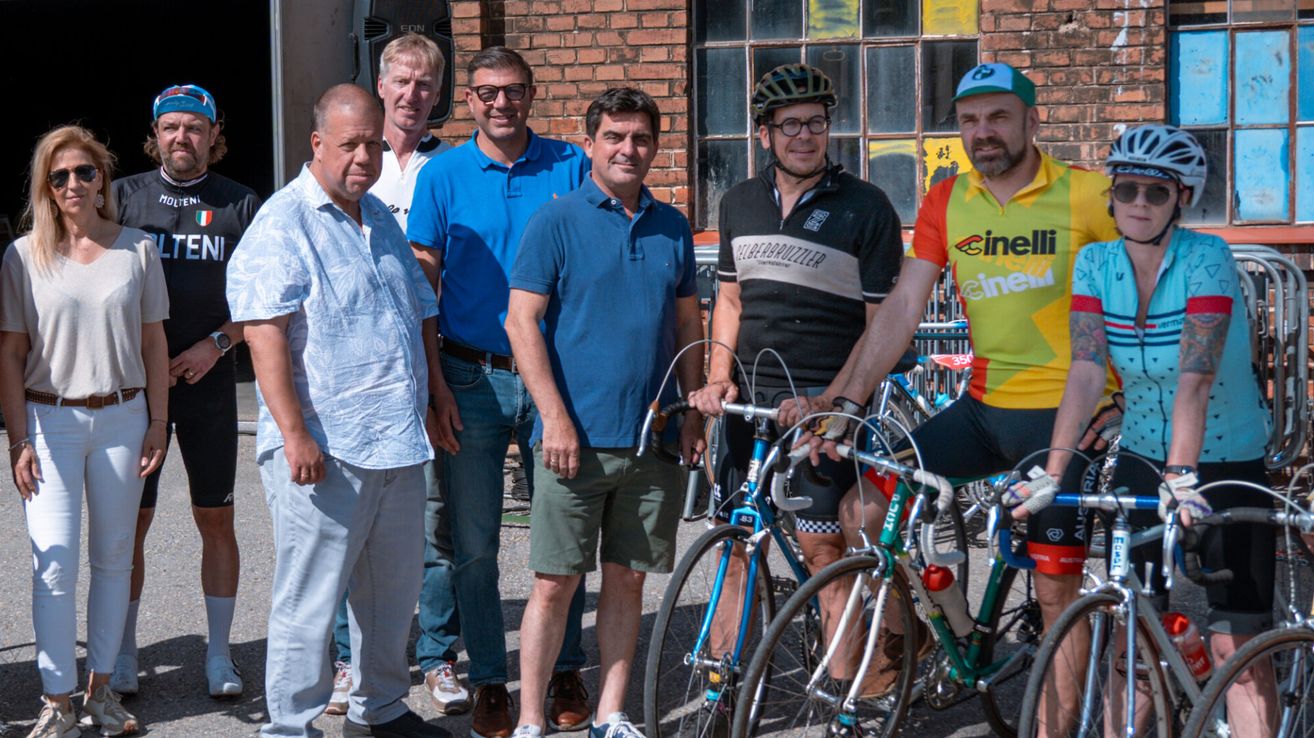 Sechs Männer stehen neben einer Ziegelwand mit zwei Fahrrädern. Ein Mann trägt Helm und Brille. Ein anderer hält ein Fahrrad, während andere Turnschuhe tragen.