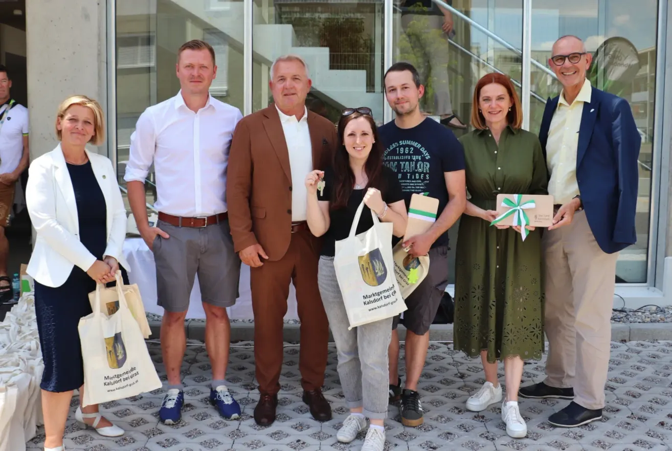 Eine Gruppe Erwachsener steht zusammen auf einem gefliesten Boden. Sie lächeln und posieren für ein Foto. Sie alle halten verschiedene Gegenstände. Eine Person hält einen Schlüssel.