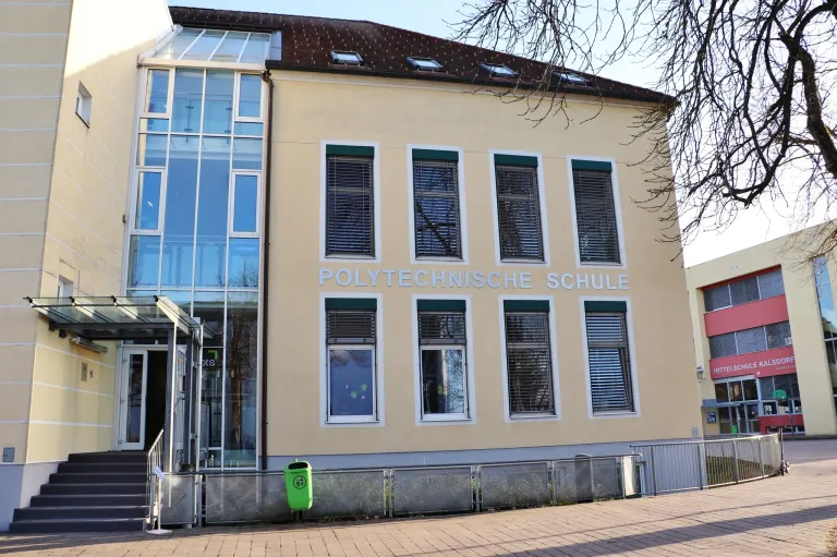 Ein beiges Gebäude mit Glasfenstern und einem Schild mit der Aufschrift 'POLYTECHNISCHE SCHUL'. Vor dem Gebäude befindet sich ein Mülleimer.