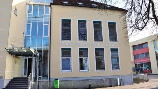 Ein beiges Gebäude mit Glasfenstern und einem Schild mit der Aufschrift 'POLYTECHNISCHE SCHUL'. Vor dem Gebäude befindet sich ein Mülleimer.
