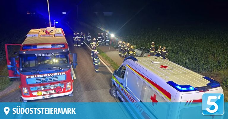 Notfallfahrzeuge, darunter ein Krankenwagen und ein Feuerwehrfahrzeug, stehen nachts am Straßenrand. Eine Gruppe Feuerwehrleute ist versammelt, und ein Autounfall ist am Straßenrand sichtbar.