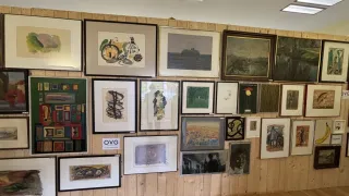 Ein Raum mit einer hölzernen Wand, geschmückt mit verschiedenen gerahmten Kunstwerken, einschließlich abstrakter und Landschaftsgemälde, mit den Initialen eines Künstlers auf einem Etikett.