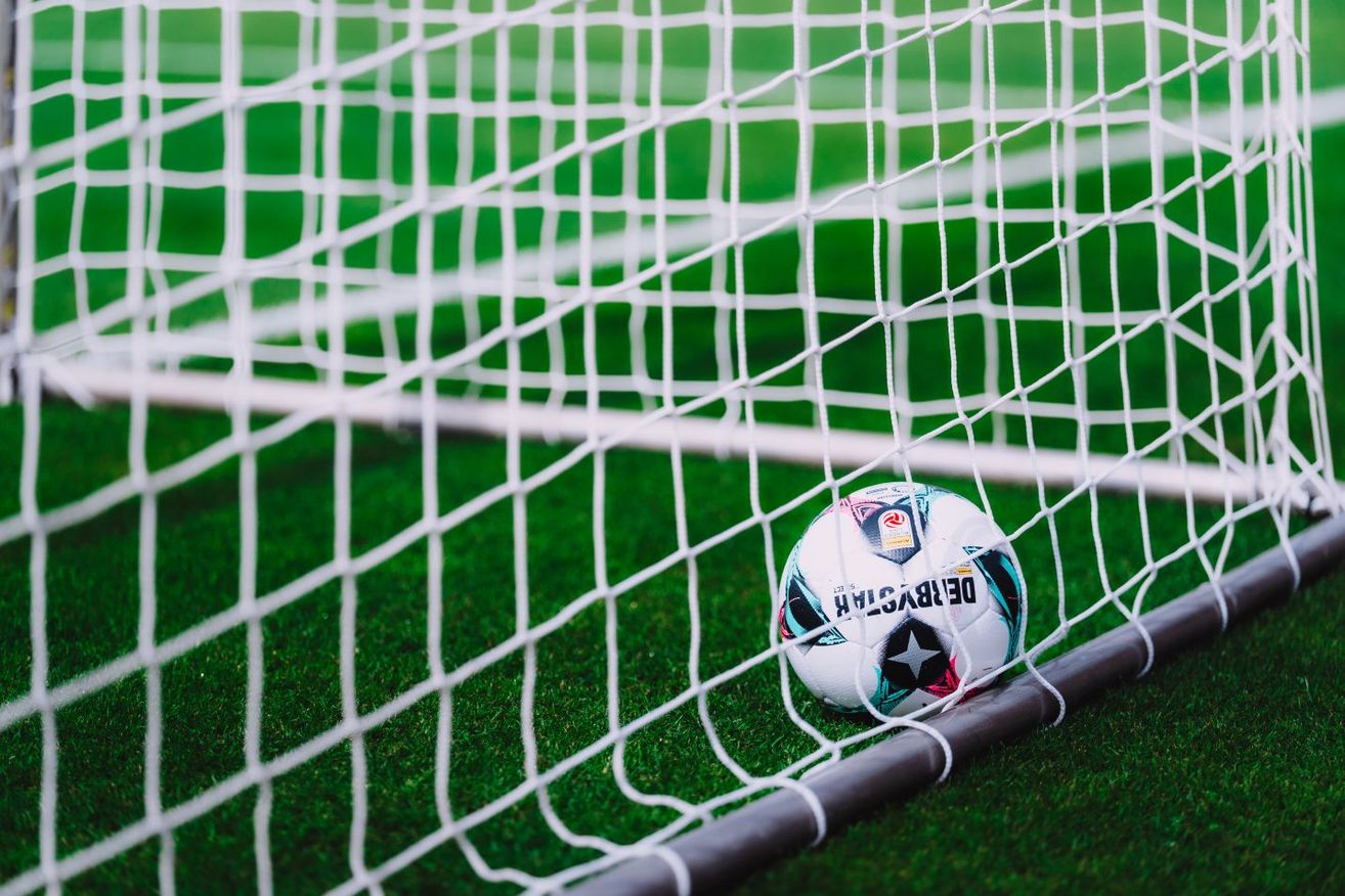 Ein Fußball liegt an einem weißen Tornetz auf einem grünen Feld. Der Ball hat farbenfrohe Designs und ein Stern-Emblem.
