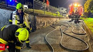 Feuerwehrleute arbeiten an einem nächtlichen Schauplatz. Einer beugt sich vor und inspiziert, während ein anderer hinter einem Zaun steht, mit einem Feuerwehrwagen in der Nähe.