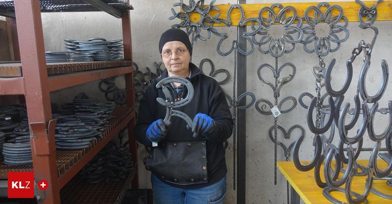 Eine Frau in einer Werkstatt hält ein Hufeisen und trägt eine Brille und Handschuhe. Hinter ihr befinden sich Regale mit vielen Hufeisen, und die Wand ist mit kunstvollen Hufeisenkunstwerken geschmückt.