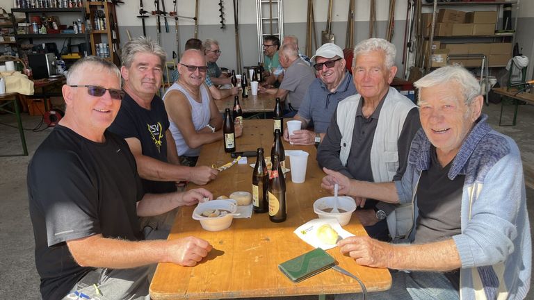Eine Gruppe älterer Männer versammelt sich um einen Tisch, lächelt und posiert für ein Foto. Sie genießen ihr Essen und Getränke. Der Tisch ist mit Flaschen, Bechern und Tellern gedeckt. Hinter ihnen stehen verschiedene Werkzeuge und Ausrüstung.